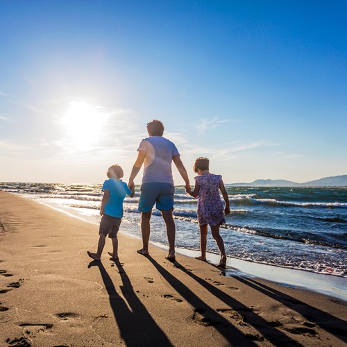 Father walking with son and daughter on a sandy beach, holding hands, rear view, lens flare